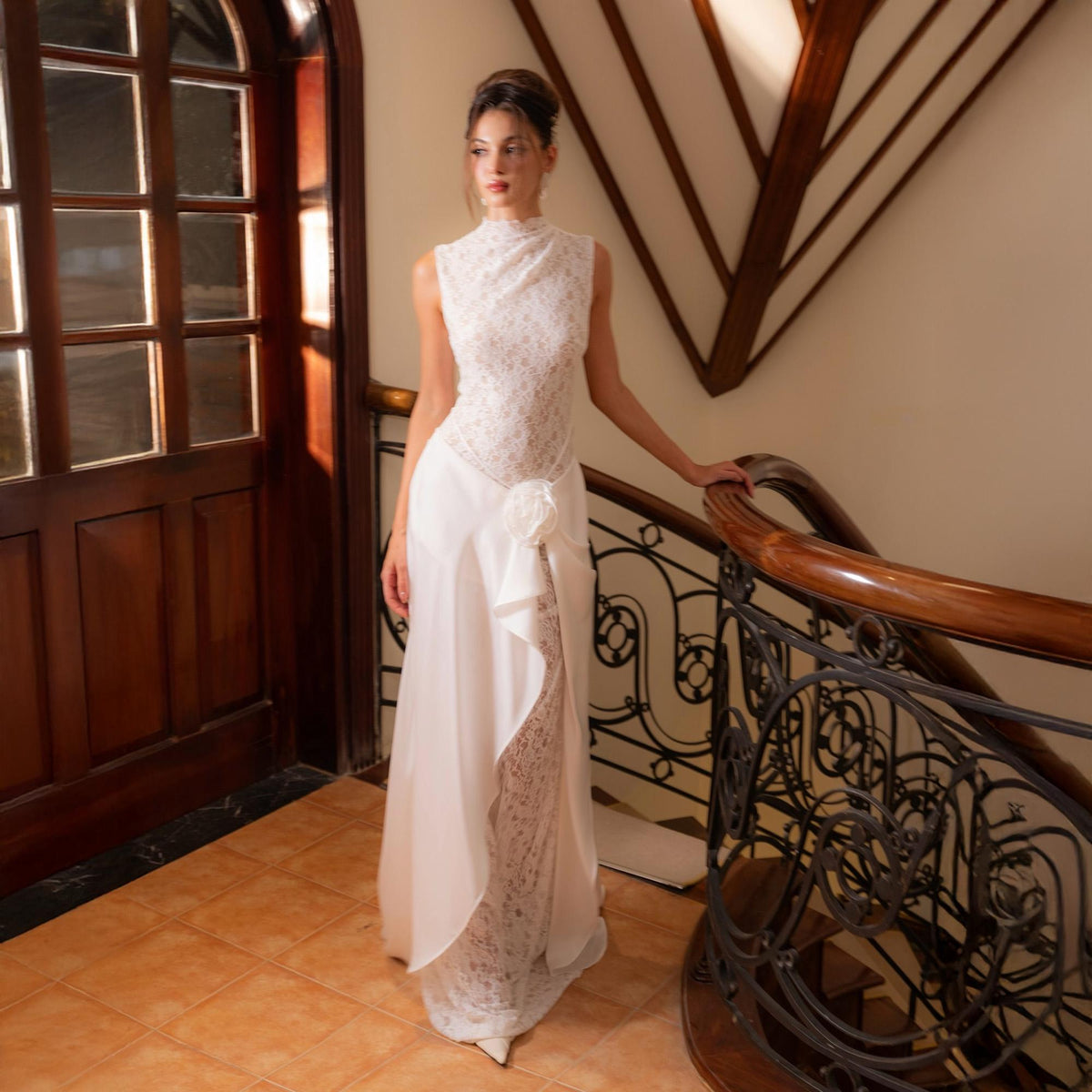May include: A white, sleeveless wedding gown with a high neckline and a flowing skirt. The bodice is adorned with delicate lace, and a large fabric flower accents the waist. The dress is worn on a staircase with a wooden banister.