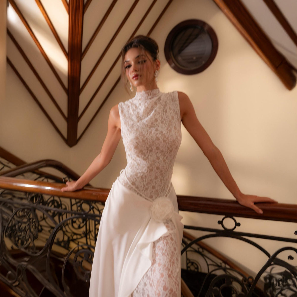 May include: A white lace wedding dress with a high neckline and a flowing skirt. The dress features a floral accent at the waist and is worn on a staircase with a wooden handrail.