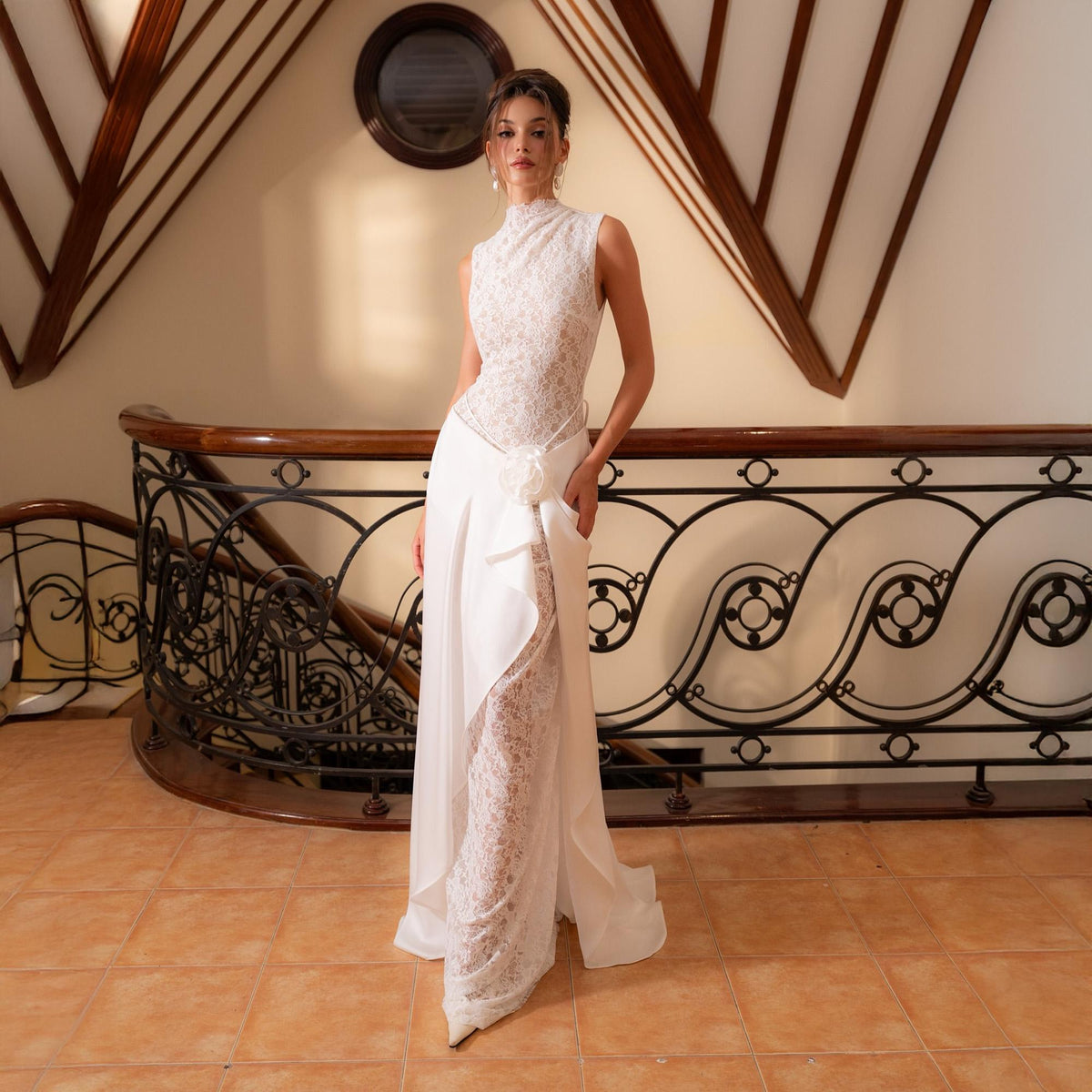 May include: A sleeveless white wedding dress with a high neckline. The bodice is made of lace, and the skirt is a flowing design with a side drape. A white flower detail accents the waist. The dress is floor-length.