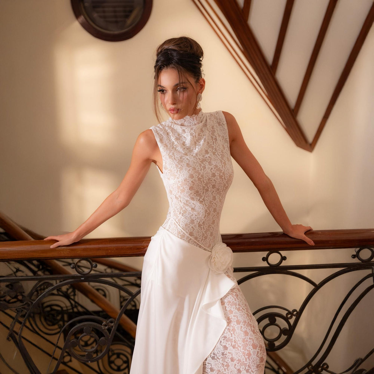 May include: A white lace bodice sleeveless dress with a high neckline and a flowing skirt. The dress features a floral accent at the waist and a lace overlay on the skirt. The model is standing on a staircase with a wooden handrail.
