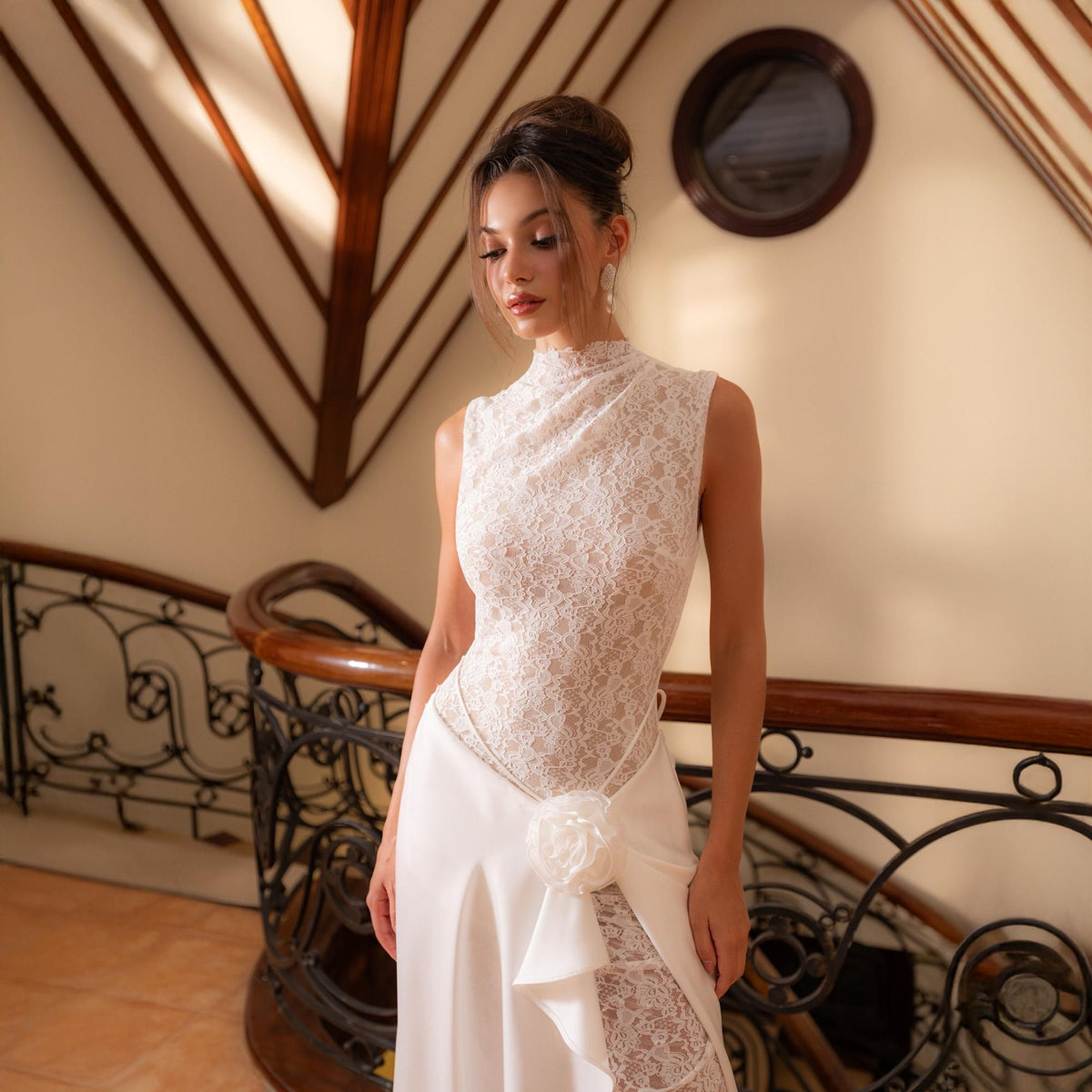 May include: A sleeveless ivory lace bodice dress with a high neckline and a flowing skirt. The dress features a floral accent at the waist and a lace panel on the skirt. The background includes a wooden staircase and a decorative railing.