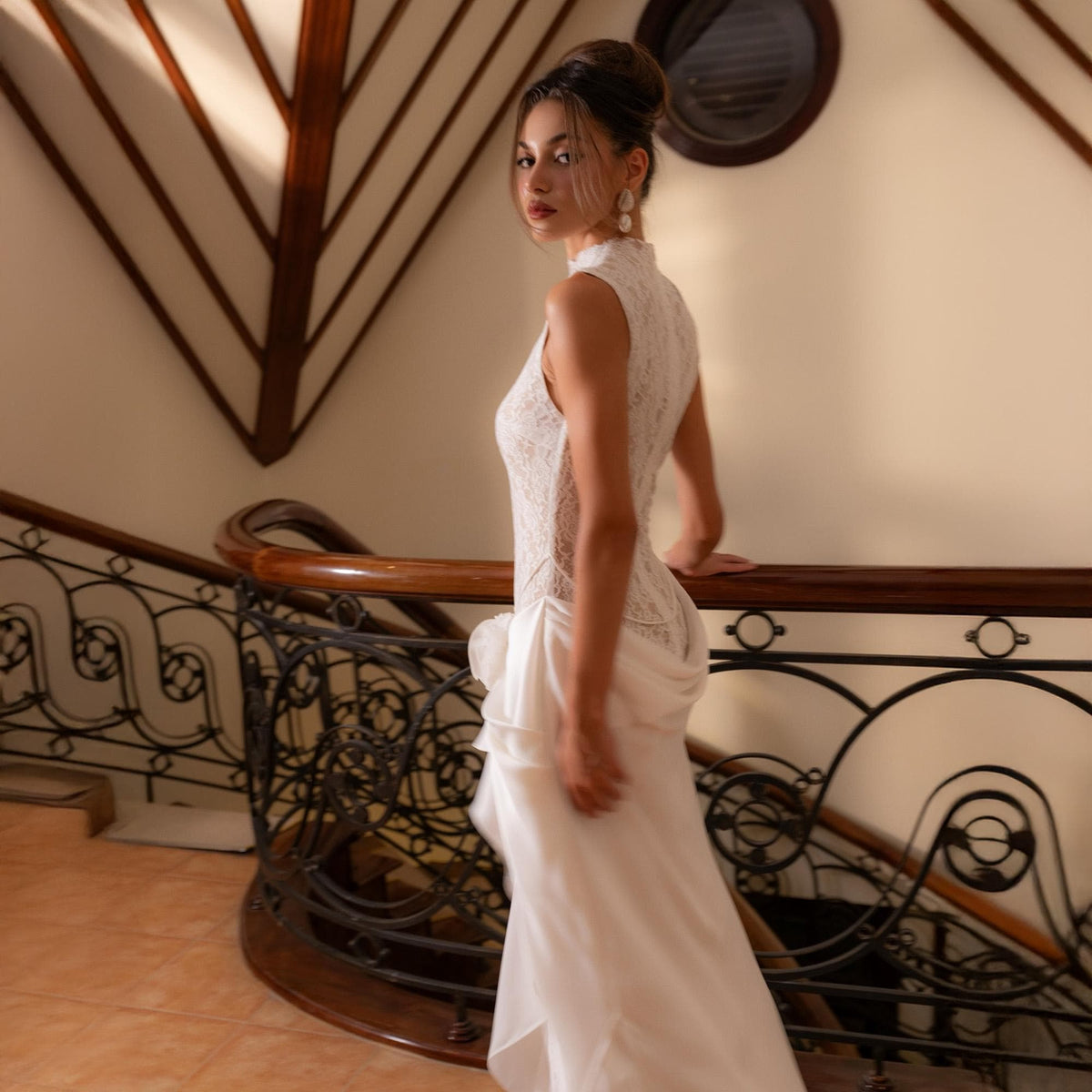 May include: A sleeveless white lace wedding dress with a high neckline and a flowing skirt. The dress features intricate lace detailing on the bodice and a layered skirt. The woman is standing on a staircase with a wooden handrail.