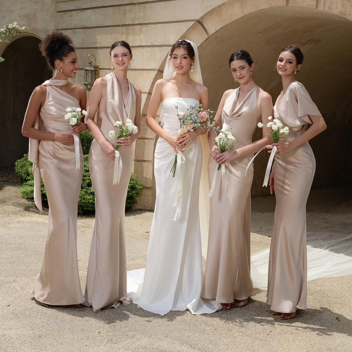 May include: A group of five people in elegant formal gowns. The bride wears a white strapless gown, while the bridesmaids wear champagne-colored dresses. Each person holds a bouquet of white flowers, and they are standing in front of an arched stone structure.