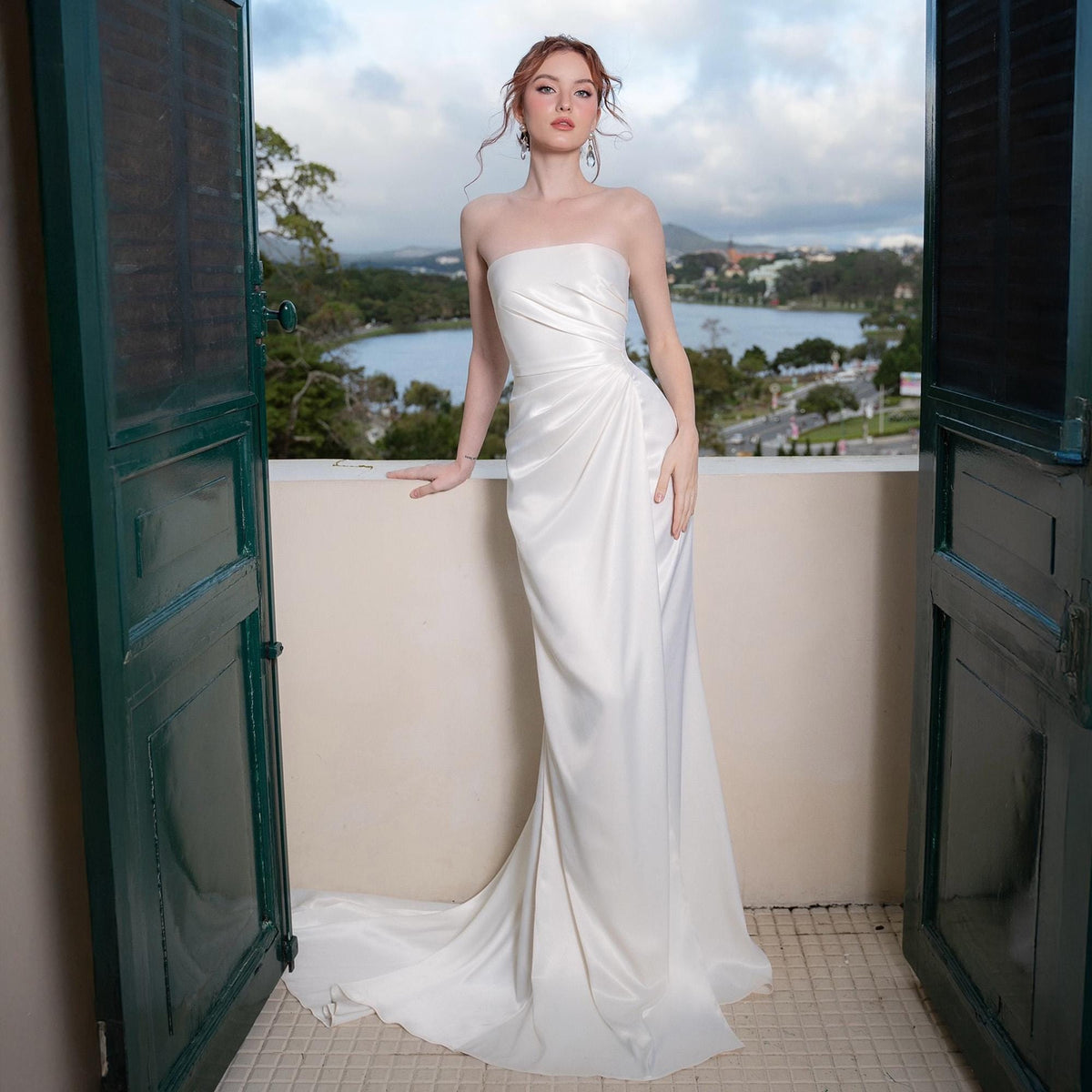 May include: A strapless, floor-length white satin gown with a draped bodice and a flowing skirt. The dress is worn by a person standing on a balcony with a scenic view of a lake and mountains in the background. The dress has a subtle train.