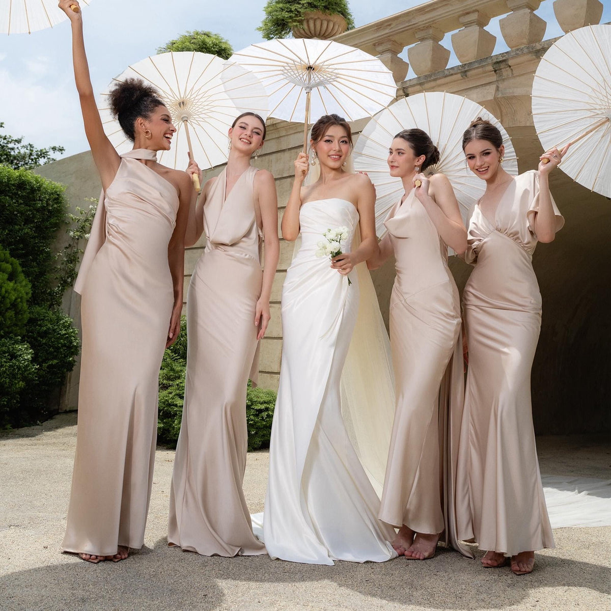 May include: Five individuals in elegant formal gowns pose outdoors, holding white parasols. The dresses are in shades of beige and white, with varying necklines and silhouettes. The scene suggests a wedding or special event.