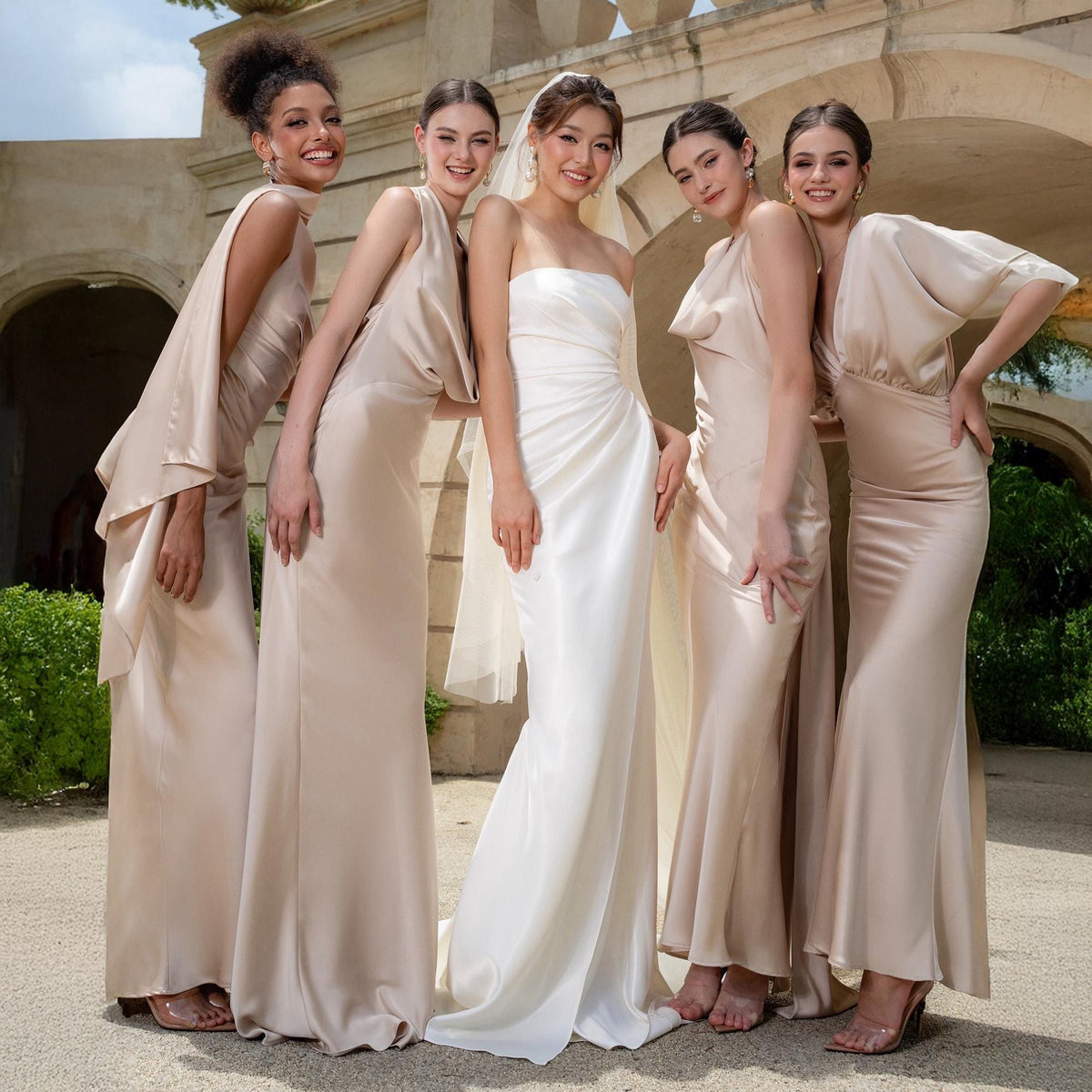 May include: Five women in elegant formal gowns pose outdoors. The bride wears a white strapless gown, while the bridesmaids wear long, flowing champagne-colored dresses with varying sleeve and neckline designs. The setting is a sunny outdoor location with architectural details.