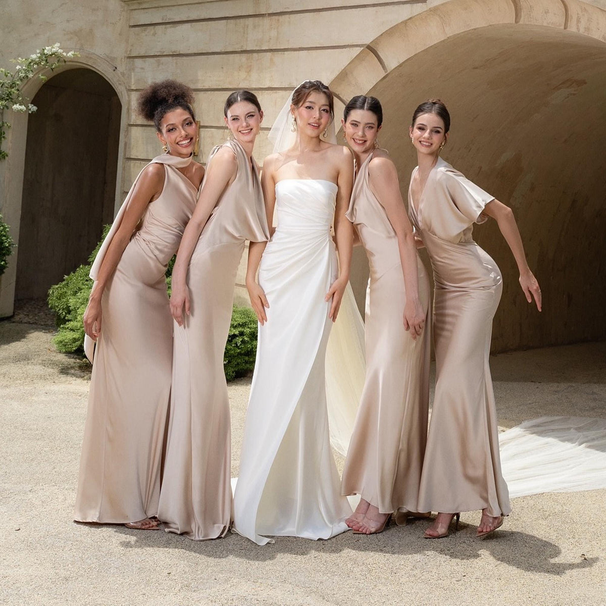 May include: A bride in a white strapless gown stands with four women in champagne-colored satin dresses. The dresses have varying necklines and sleeve styles. The group is posed outdoors in front of a stone archway.