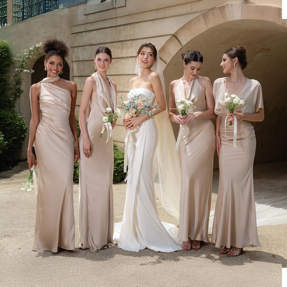 May include: Five women in elegant, floor-length gowns. The dresses are in shades of champagne and ivory, with varying necklines and styles. Each woman holds a bouquet of flowers, suggesting a wedding or formal event.