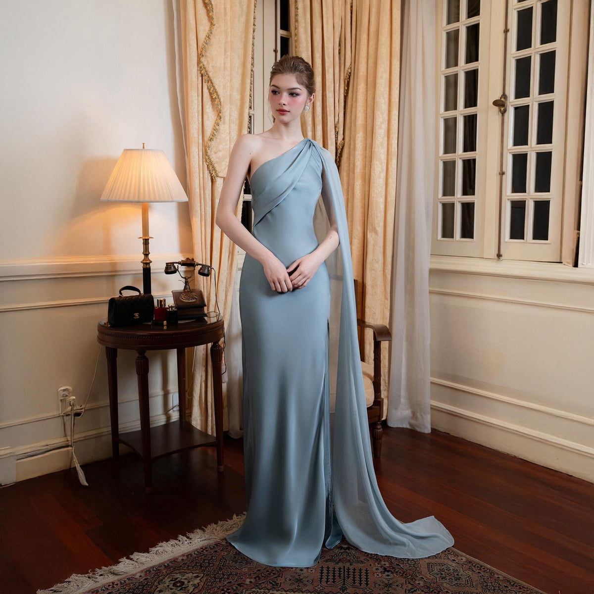 May include: A light blue, one-shoulder formal gown with a flowing cape detail. The dress has a fitted silhouette and a high slit. The model is standing in a room with a lamp, table, and curtains. The dress is floor-length.