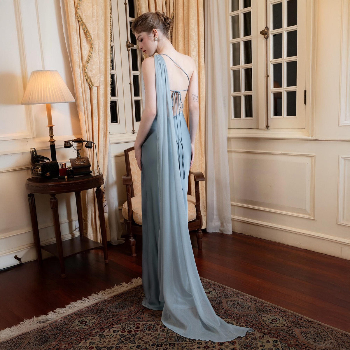 May include: A light blue, floor-length gown with a draped, flowing design. The dress features a single shoulder strap and a low back with delicate tie detailing. The gown's fabric cascades down the back, creating a dramatic train.