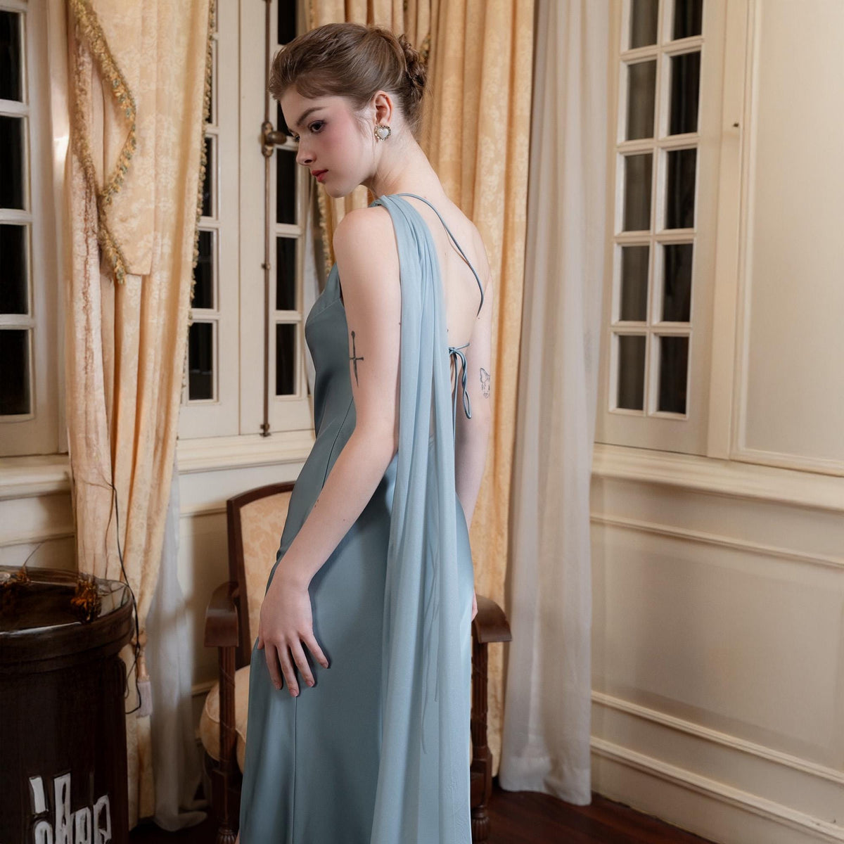 May include: A long, flowing, pale blue dress with a draped, sheer overlay. The dress features a simple, elegant design with spaghetti straps and a low back. The fabric appears to be a soft, lightweight material, suitable for formal occasions.