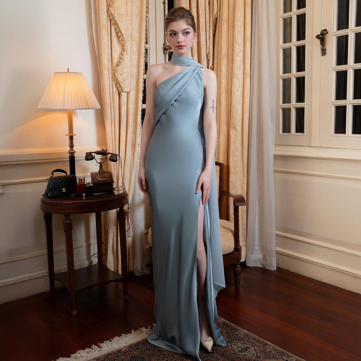 May include: A light blue, one-shoulder evening gown with a high slit. The dress features a draped neckline and a flowing, floor-length skirt. The model is standing in a room with a lamp, a table, and a window.