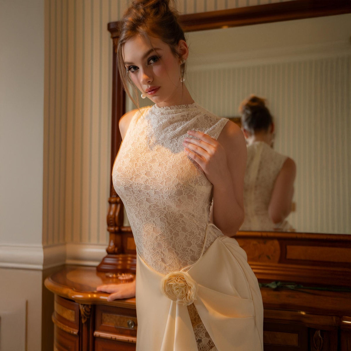 May include: A cream-colored, sleeveless wedding dress with a high, lace neckline and a flowing skirt. The dress features a floral accent at the waist and is worn in front of a wooden vanity with a mirror.