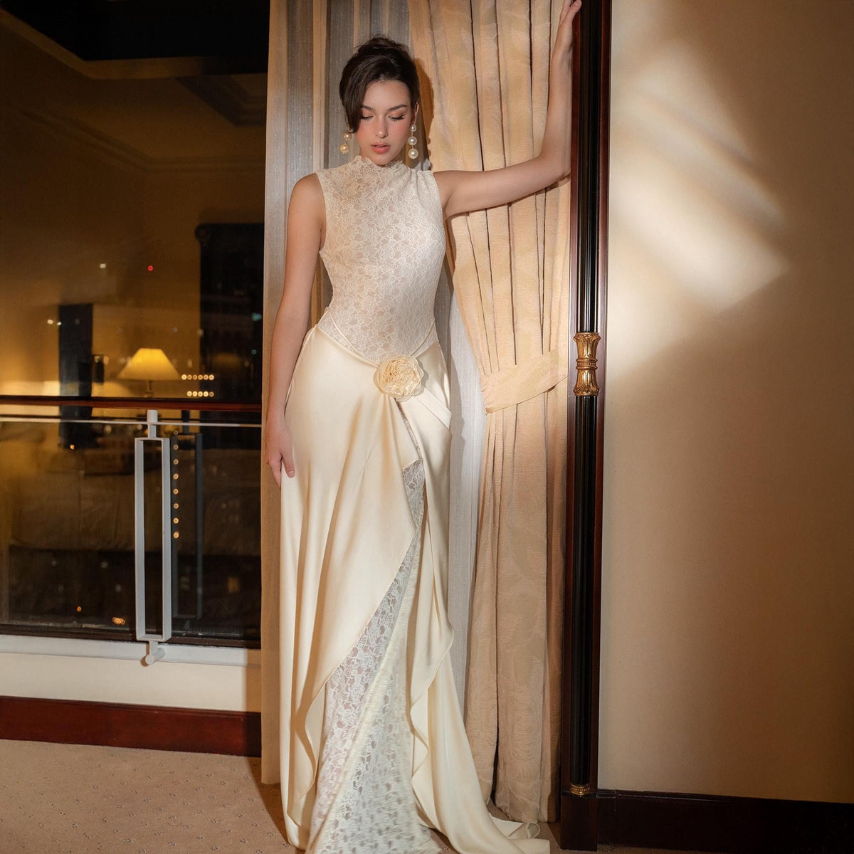 May include: A sleeveless, floor-length ivory gown with a high neckline and a floral accent at the waist. The bodice features a lace overlay, and the skirt has a layered design with a lace panel. The dress is suitable for formal events.