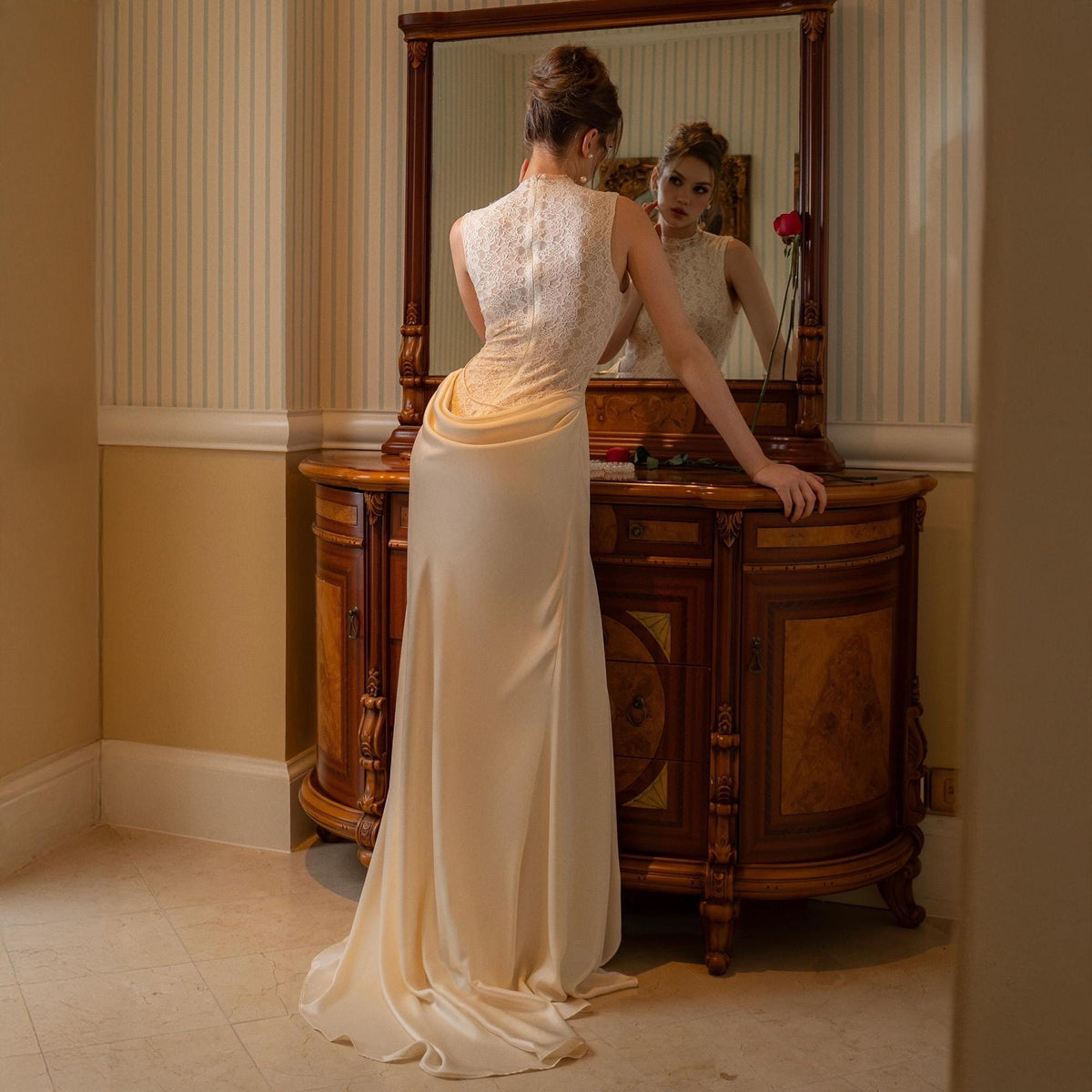 May include: A cream-colored, sleeveless gown with a high neckline and a flowing skirt. The bodice features delicate lace detailing, and the dress has a subtle train. The woman is standing in front of a large wooden mirror.