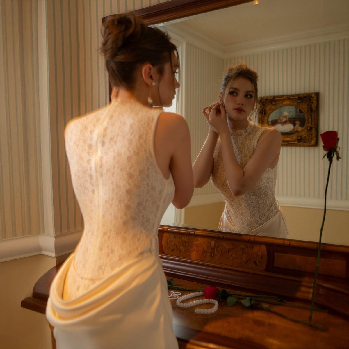 May include: A woman in a white lace dress is reflected in a mirror, adjusting an earring. A red rose and a pearl necklace rest on the wooden vanity. The room has a vintage aesthetic with striped wallpaper and a framed painting.