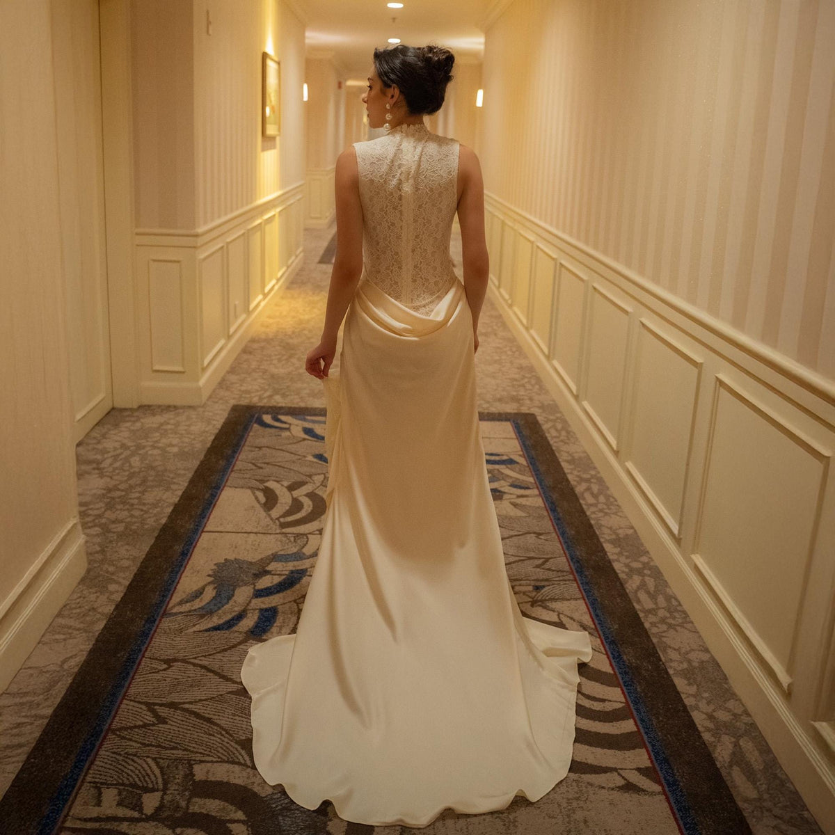 May include: A woman in a cream-colored wedding gown with a long train walks down a hallway. The dress features a high neckline and a detailed lace back. The hallway has striped wallpaper and a patterned carpet.