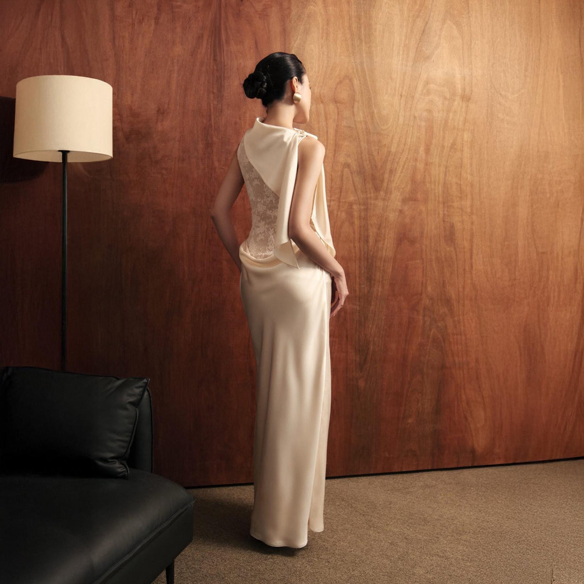 May include: A cream-colored, floor-length gown with a draped, cape-like overlay. The back of the dress features a lace panel. The model is standing in front of a wood-paneled wall. A black leather sofa and a floor lamp with a beige lampshade are in the background.