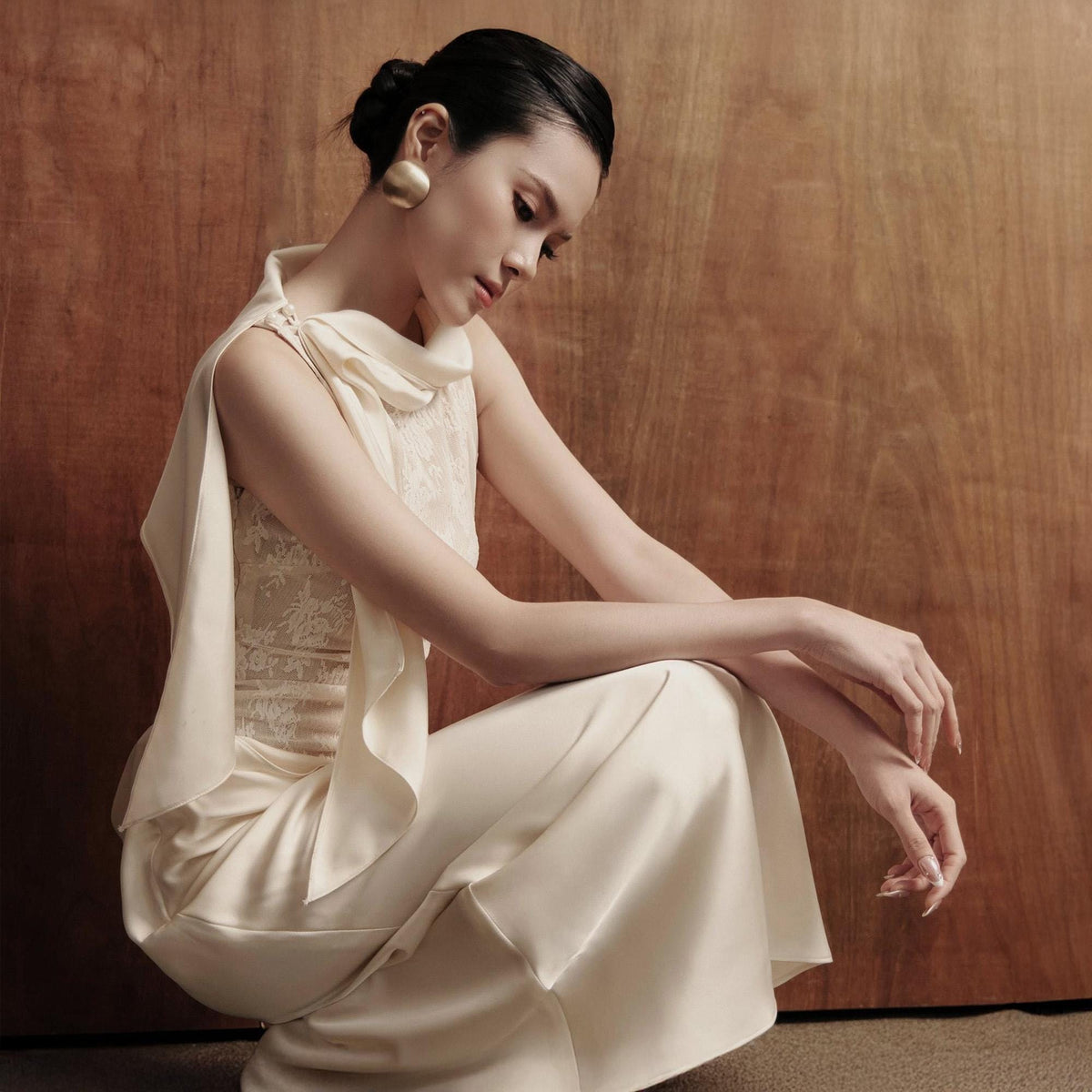 May include: An ivory-colored formal dress with a draped cowl neckline and a flowing skirt. The dress features a lace bodice and is paired with large gold earrings. The model is seated in front of a wooden backdrop.
