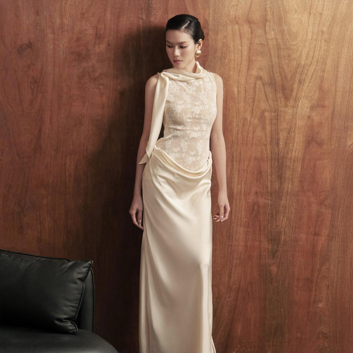May include: A sleeveless, floor-length ivory gown with a draped cowl neckline and a fitted bodice. The bodice features a delicate lace overlay, and the skirt flows smoothly. The model is standing in front of a warm wood-paneled wall.
