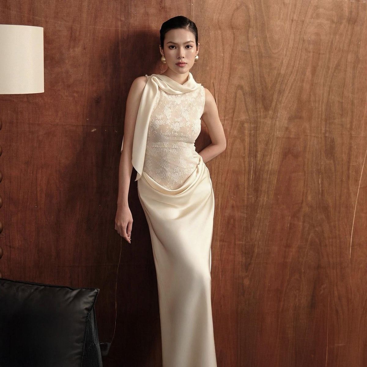 May include: A sleeveless ivory gown with a draped cowl neckline and a fitted bodice. The dress features a flowing skirt and is made of a silky, elegant fabric. The model is wearing gold earrings.