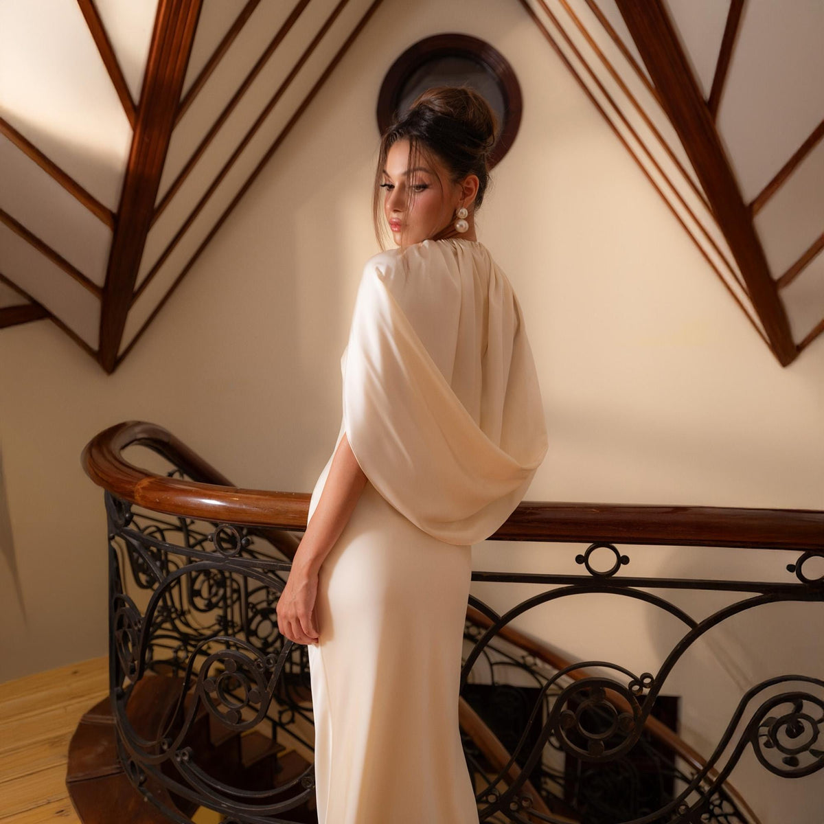 May include: A cream-colored, floor-length gown with a draped cape-like overlay. The dress is worn on a wooden staircase with a decorative wrought iron railing. The model is wearing pearl earrings and has her hair up.
