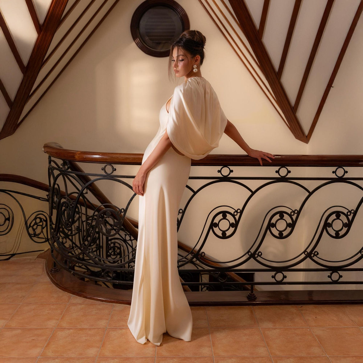 May include: A cream-colored, floor-length gown with a draped, cape-like overlay. The dress features a simple, elegant design, perfect for formal events. The model is standing on a staircase with a decorative railing.