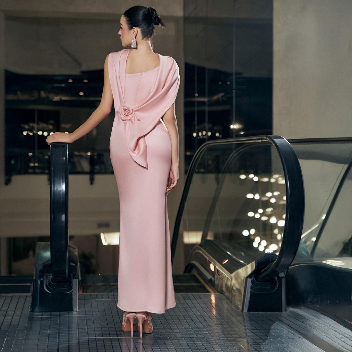 May include: A light pink, floor-length gown with a draped back and a rose detail. The dress features a square neckline and a fitted silhouette. The woman is wearing matching pink heels and earrings. The background includes an escalator and city lights.