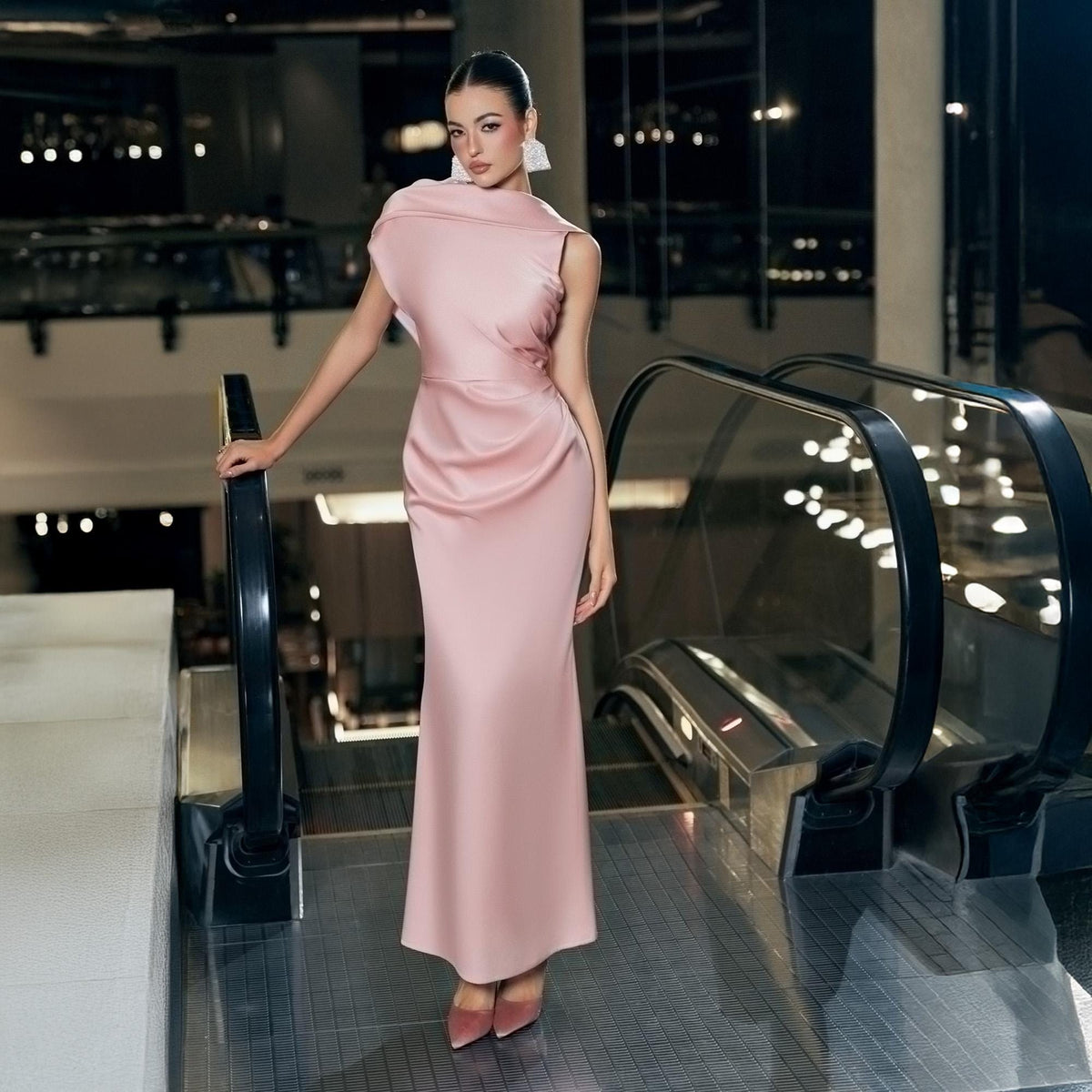 May include: A sleeveless, floor-length, blush pink satin gown. The dress features a draped neckline and a fitted silhouette. The model is wearing matching pink heels. The background includes an escalator and ambient lighting.