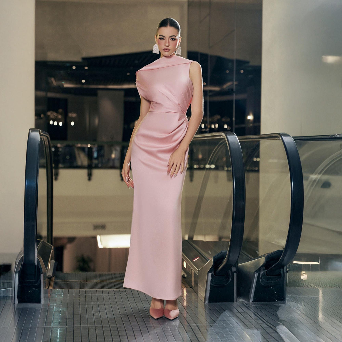 May include: A sleeveless, floor-length, blush pink gown with a high neckline and draped bodice. The dress features a fitted waist and a straight skirt. The model is wearing matching pink heels and large white earrings. The background includes an escalator.