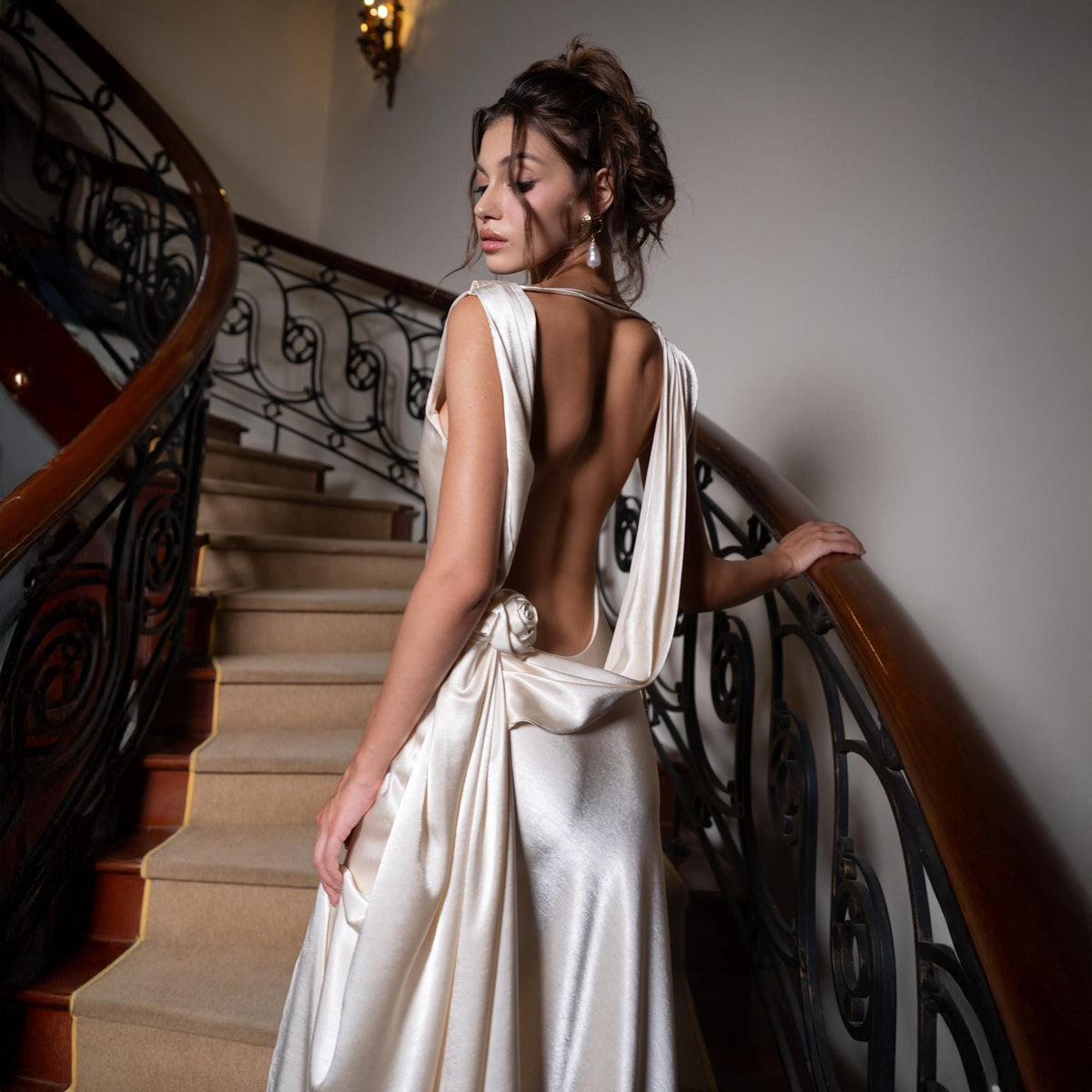 May include: A cream-colored, backless formal gown with a draped design. The dress features a low back and a small floral accent at the waist. The model is standing on a staircase with a wooden handrail.
