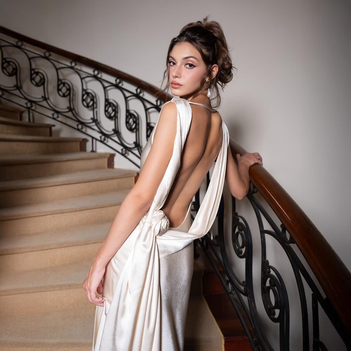 May include: A cream-colored, backless formal gown with a draped design. The dress features a flowing silhouette and is worn on a staircase with a dark wooden handrail. The model is looking over their shoulder.