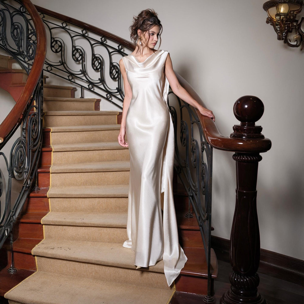 May include: A floor-length ivory satin gown with a cowl neckline and a flowing, open back design. The dress is sleeveless and drapes elegantly. The model is standing on a carpeted staircase with a wooden banister.