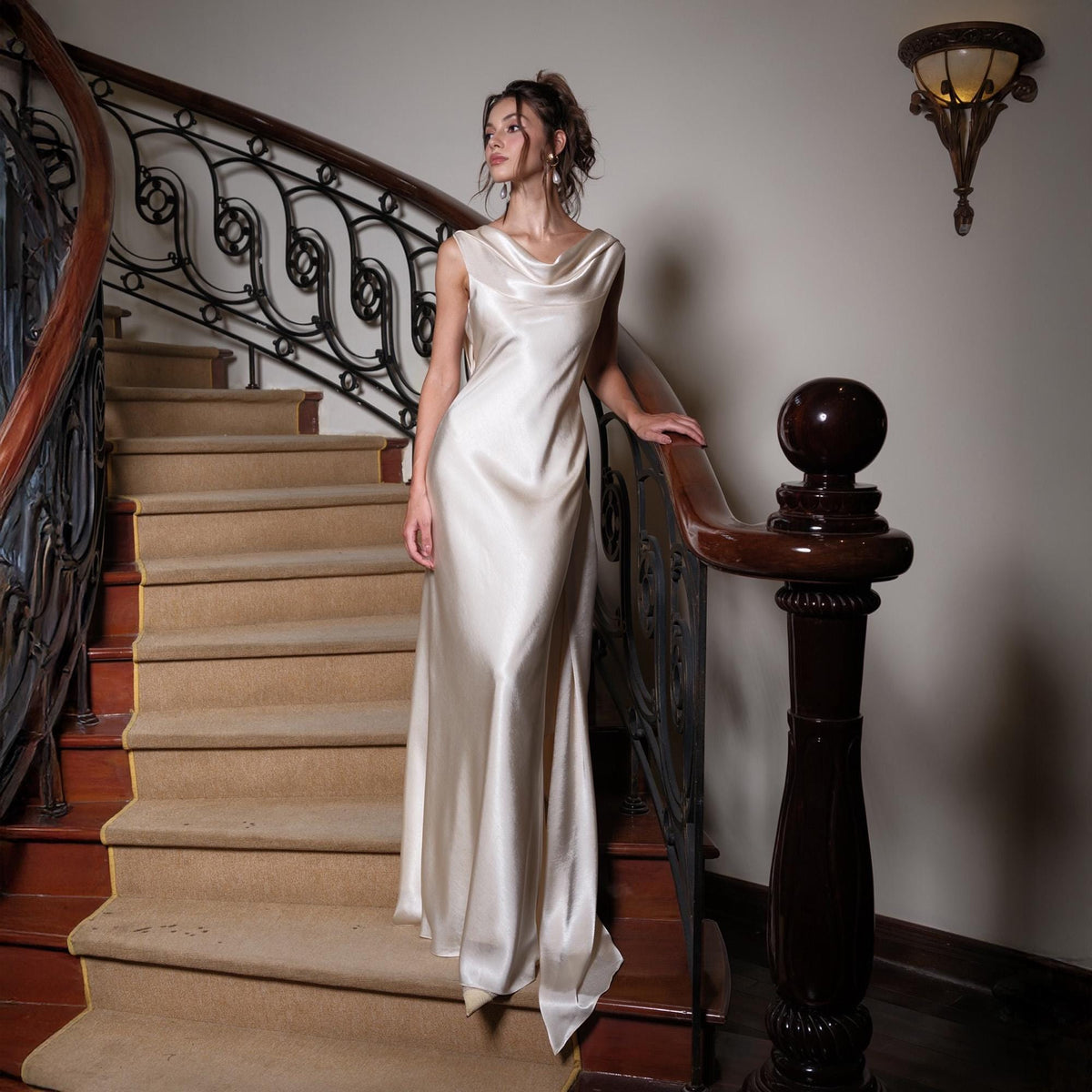 May include: A cream-colored, sleeveless satin gown with a cowl neckline. The dress is floor-length and drapes elegantly. The image shows the dress on a staircase with a dark wood banister and beige carpeted steps.