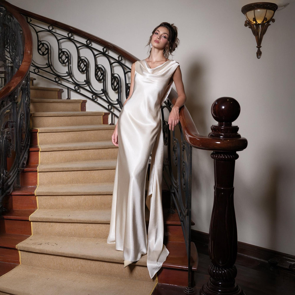 May include: A floor-length, ivory satin gown with a cowl neckline and a flowing skirt. The dress is worn on a staircase with a dark wood banister and ornate wrought iron railing. The stairs are covered in a neutral-toned carpet.
