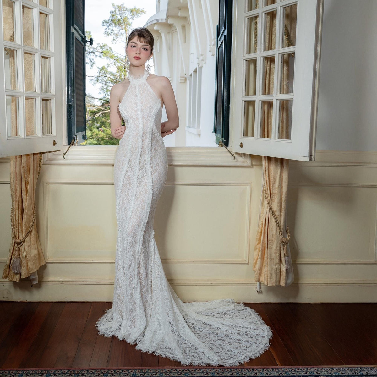 May include: A white lace halter-neck gown with a mermaid silhouette and a long train. The dress features intricate floral lace detailing and a fitted bodice. The gown is worn in front of a window with natural light.