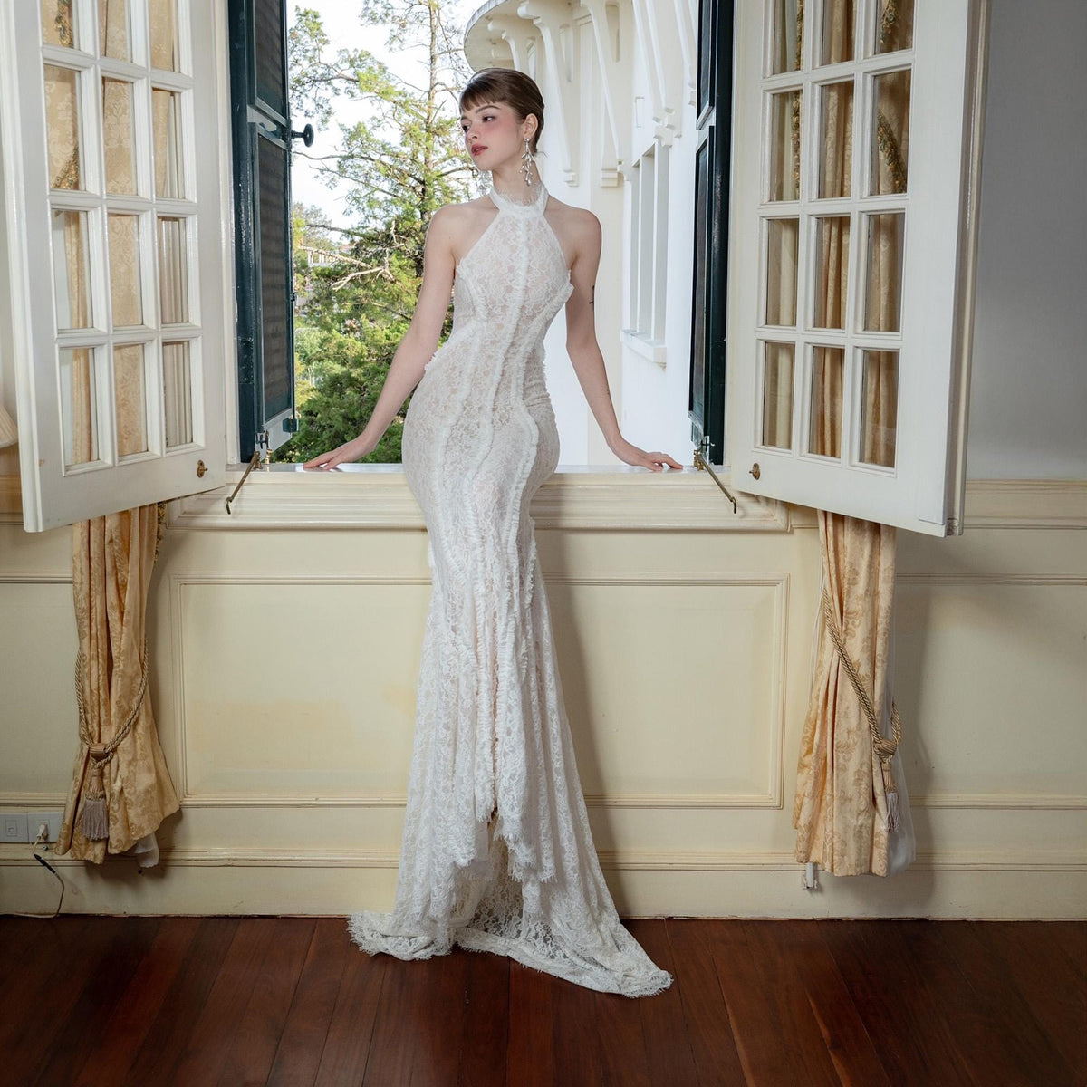 May include: A white, floor-length halter dress with a delicate lace overlay. The dress features a fitted bodice and a flowing skirt with a slight train. The model is standing near an open window with a view of trees.