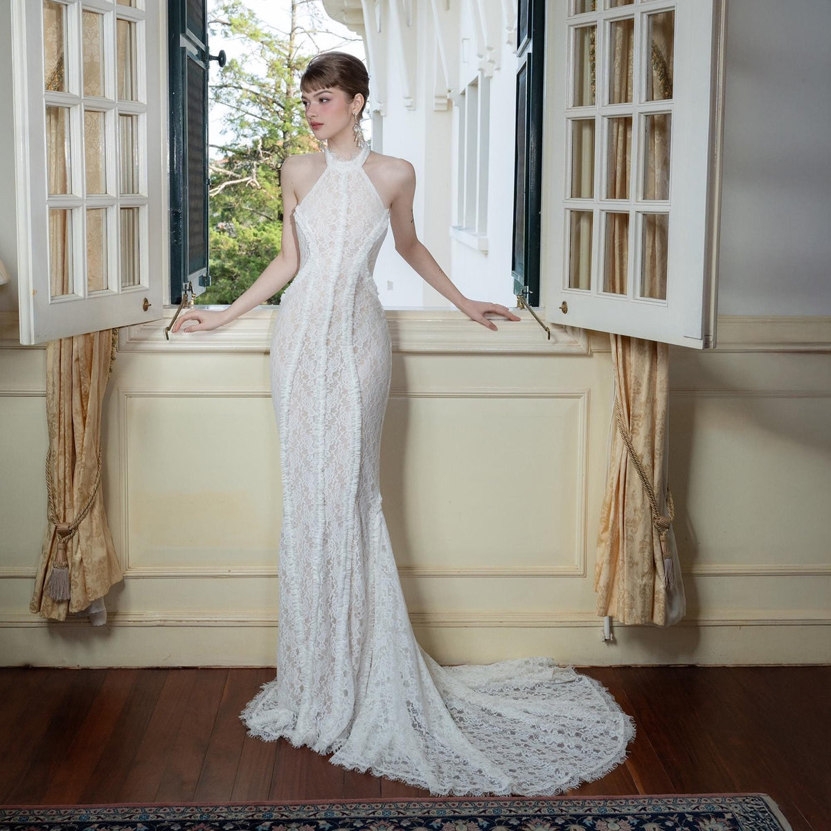 May include: A white lace halter neck gown with a mermaid silhouette and a long train. The dress features a high neckline and intricate lace detailing. The gown is displayed near an open window with natural light.