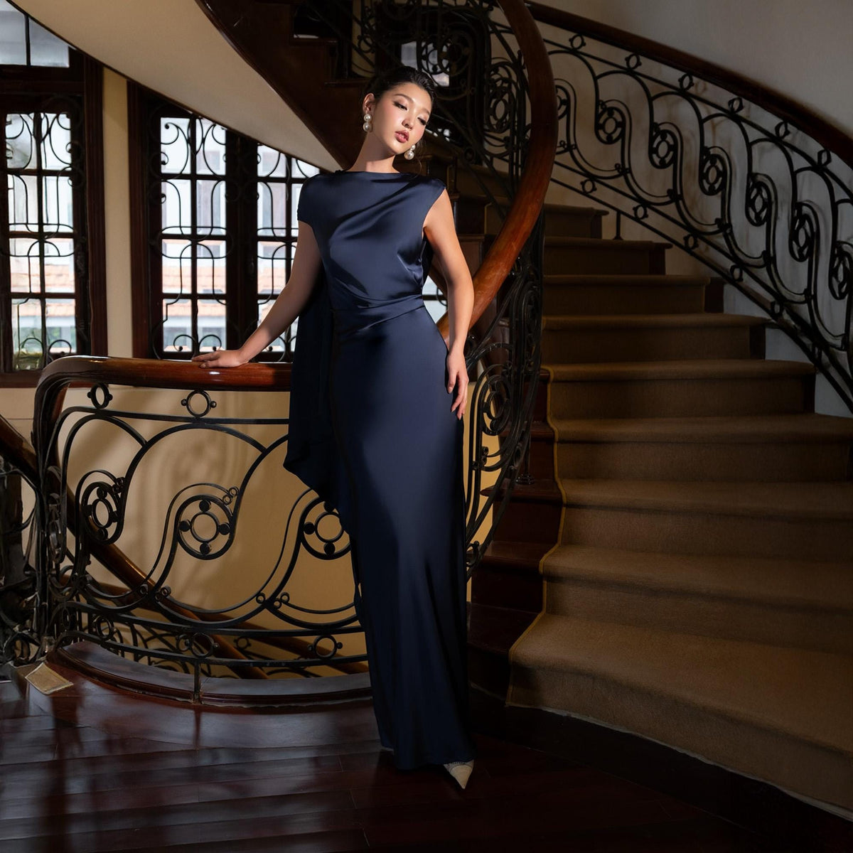 May include: A navy blue, floor-length gown with a draped, asymmetrical design. The dress features a boat neckline and cap sleeves. The woman is standing on a wooden staircase with a decorative wrought iron railing.