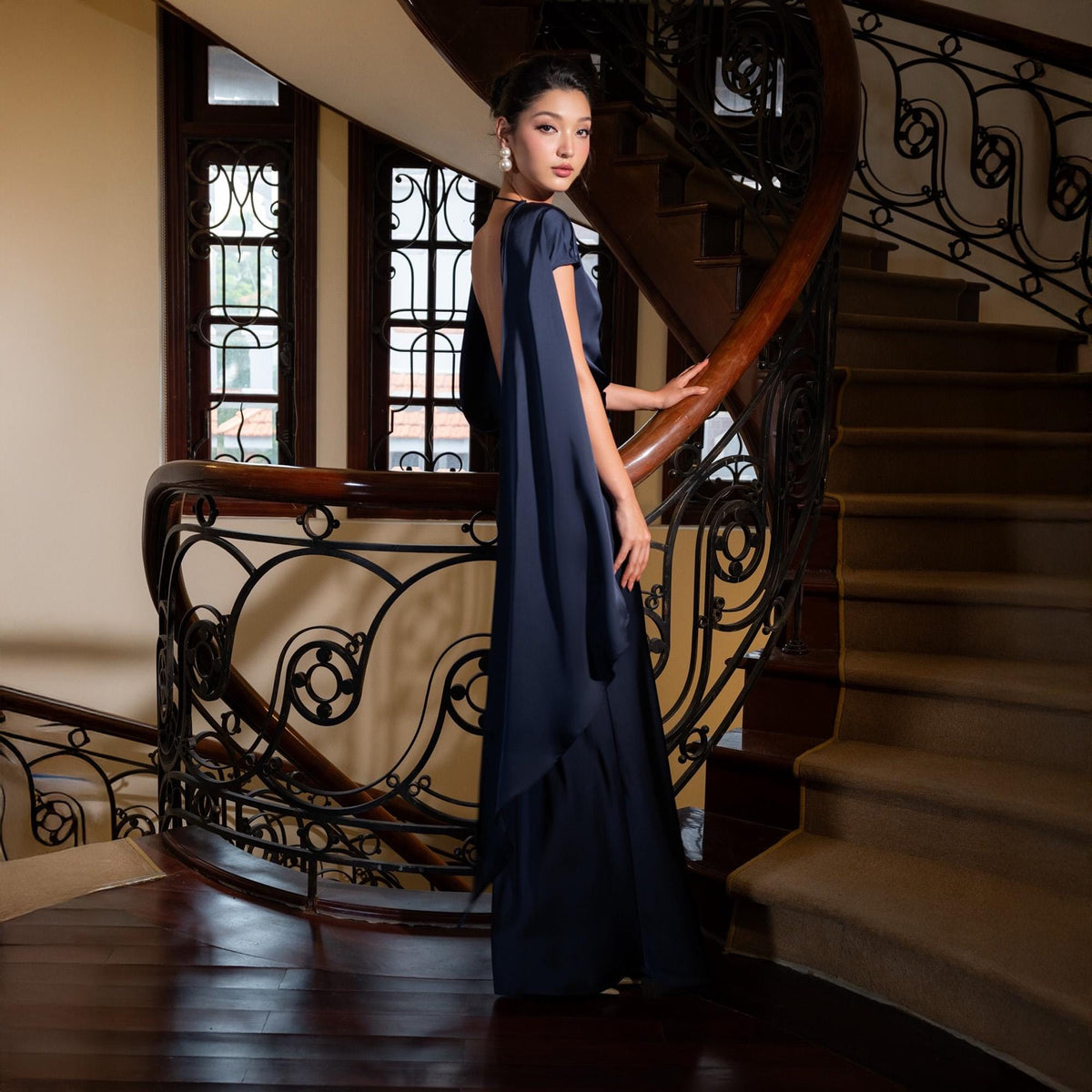 May include: A navy blue, floor-length gown with a cape-like design. The dress features a low back and short sleeves. The model is standing on a wooden staircase with ornate wrought iron railings.