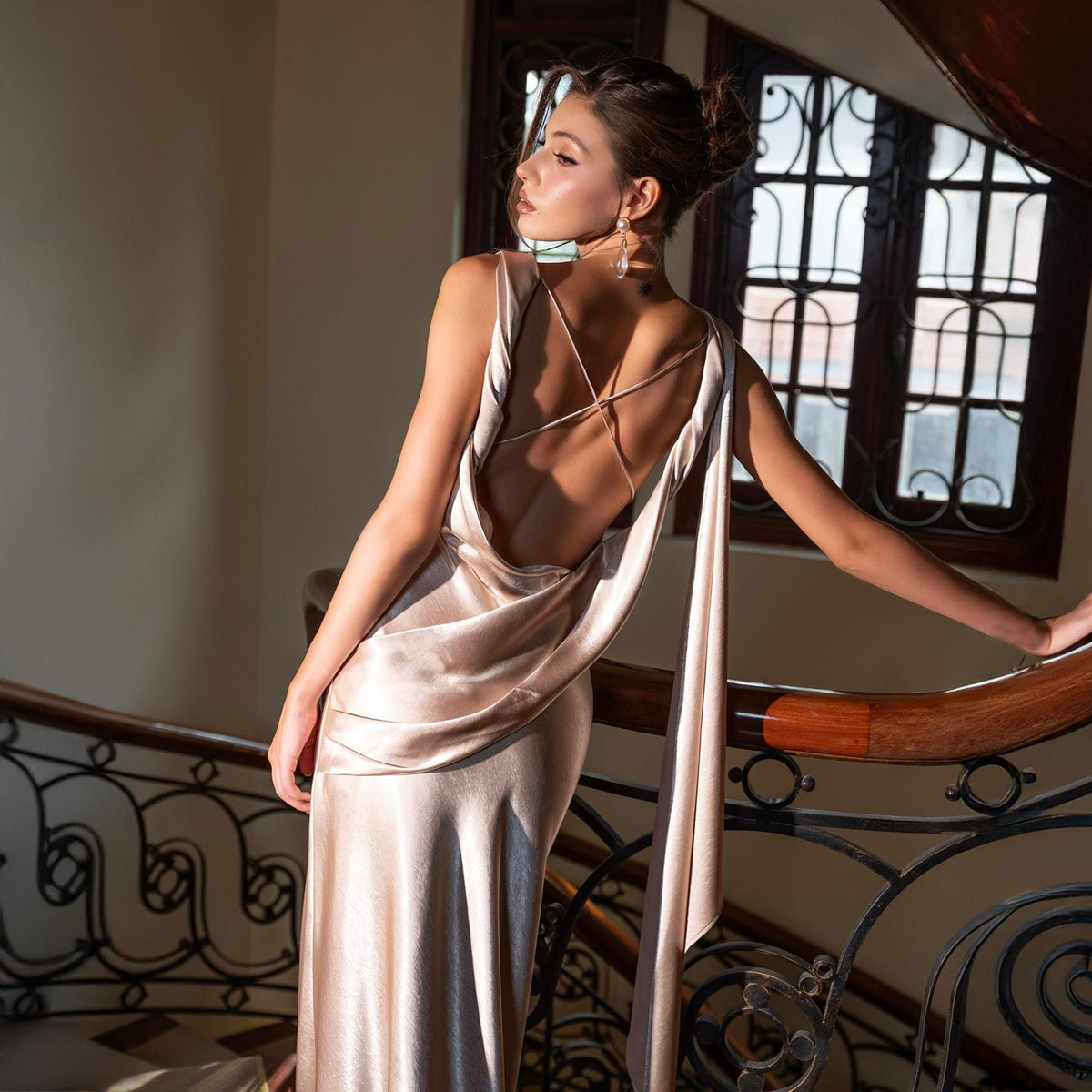 May include: A champagne-colored satin gown with a low back and draped cowl neckline. The dress features delicate spaghetti straps that crisscross in the back and a flowing, floor-length skirt. The model is standing on a wooden staircase with a wrought iron railing.