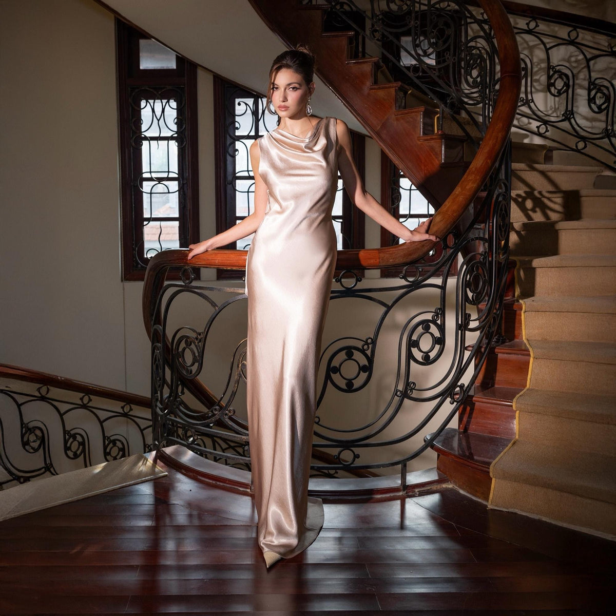May include: A floor-length, sleeveless, champagne-colored satin gown. The dress features a cowl neckline and a form-fitting silhouette. The woman is standing on a wooden staircase with ornate wrought iron railings.