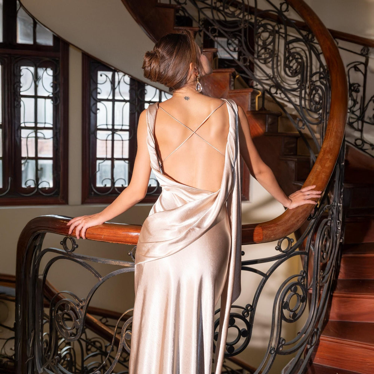 May include: A champagne-colored, backless evening gown with delicate spaghetti straps that crisscross the back. The dress drapes elegantly, creating a flowing silhouette. The model is standing on a wooden staircase with ornate metal railings.