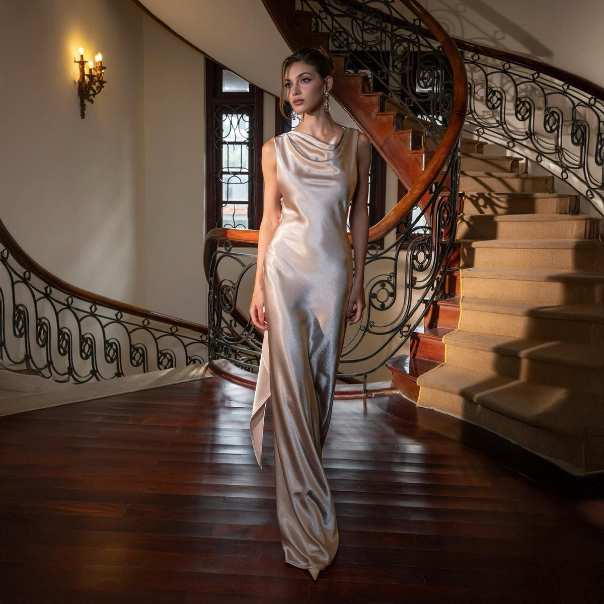 May include: A floor-length, champagne-colored satin gown with a cowl neckline. The dress has a flowing silhouette and a subtle sheen, perfect for formal events. The background features a grand staircase with a dark wood banister.