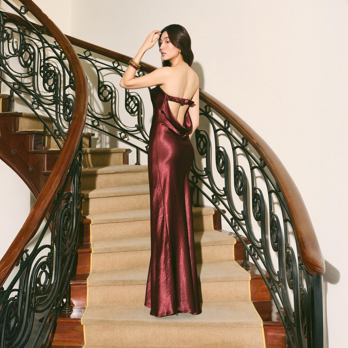 May include: A burgundy satin gown with a low back and a bow detail. The dress is floor-length and has a slight sheen. The model is standing on a carpeted staircase with a wooden banister.
