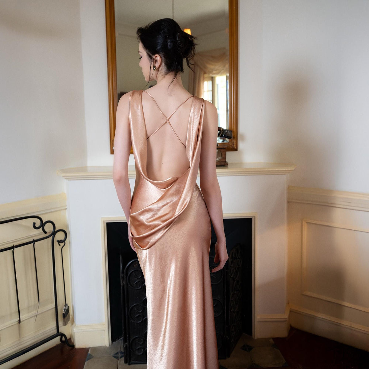May include: A shimmering, rose-gold evening gown with a cowl back and delicate crisscross straps. The floor-length dress has a flowing silhouette, perfect for formal events. The model is standing in front of a fireplace with a mirror.