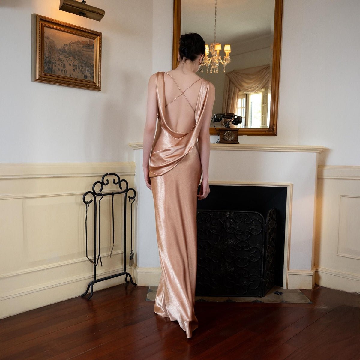 May include: A floor-length, rose-gold satin dress with a cowl neckline and crisscross back straps. The dress is worn by a person standing in a room with a fireplace, mirror, and a decorative metal stand.