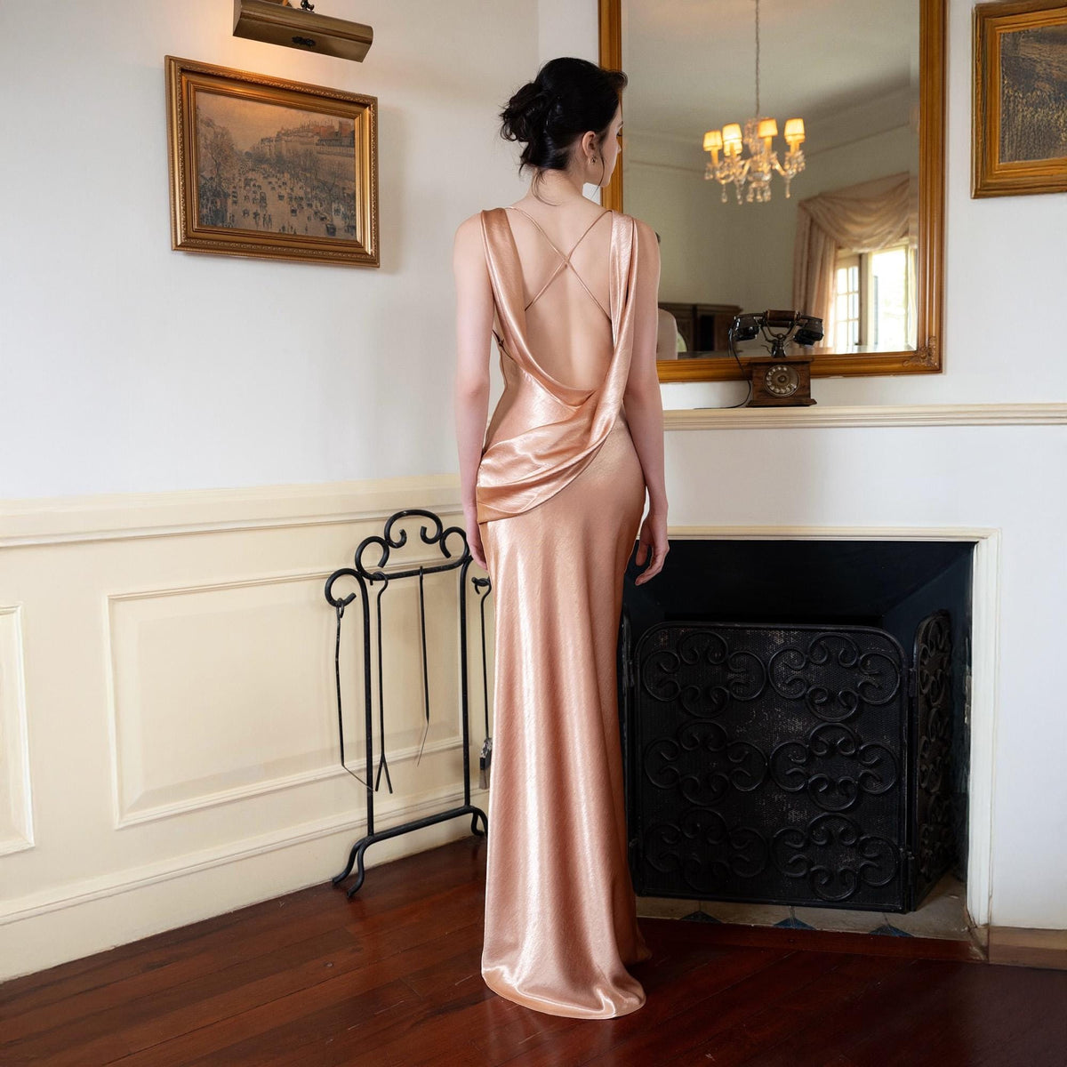 May include: A floor-length, rose-gold satin gown with a cowl back and spaghetti straps. The dress has a sleek, form-fitting silhouette and a slight train. The model is standing in front of a fireplace and mirror.