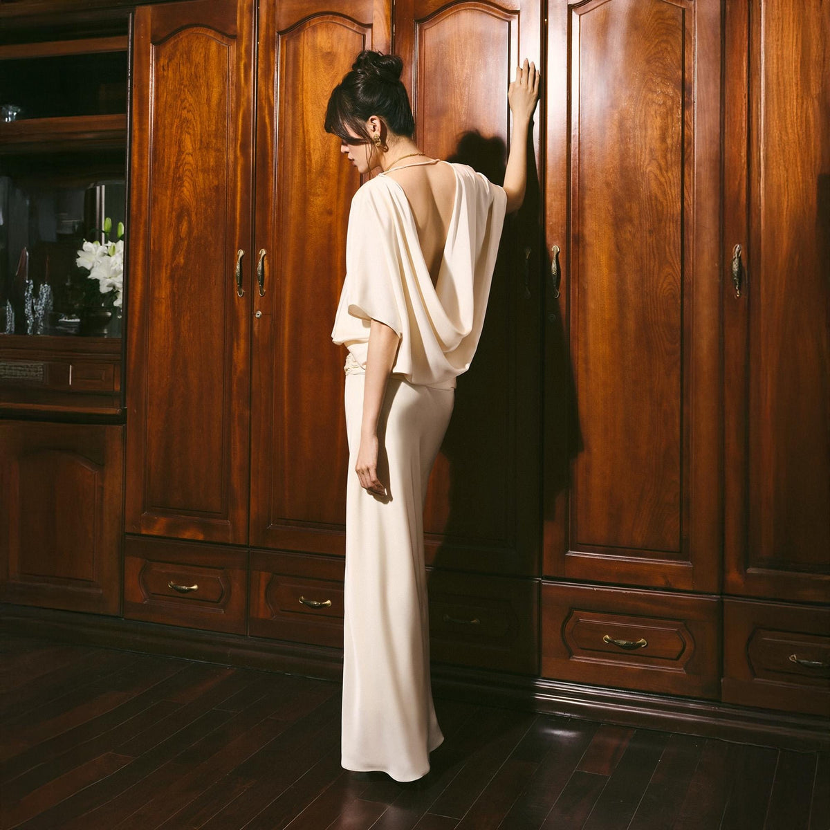 May include: A cream-colored, floor-length gown with a draped cowl back and short sleeves. The dress has a fitted waist and a flowing skirt. The model is standing in front of a dark wood wardrobe.