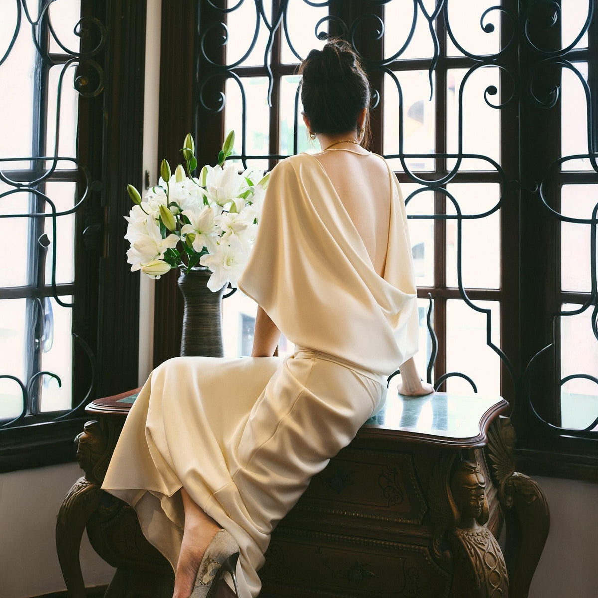 May include: A cream-colored, draped backless dress. The dress is worn by a person sitting on a dark wooden dresser. A vase of white lilies sits near a large window with decorative ironwork.